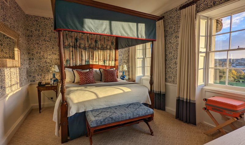 four poster bed with white sheets and dark wooden frame in a hotel bedroom wioth pale blue patterned wallpaper and a window looking out onto a green grassy park