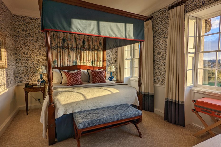 four poster bed with white sheets and dark wooden frame in a hotel bedroom wioth pale blue patterned wallpaper and a window looking out onto a green grassy park