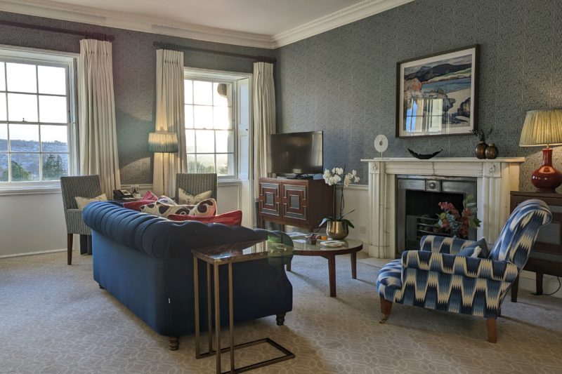 Ruang tamu yang nyaman dengan dinding biru, karpet krem, dan sofa biru menghadap perapian dengan panel krem.  Ulasan tentang Royal Crescent Hotel dan Pemandian Spa. 