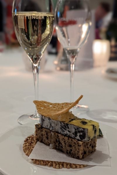 two champagne glasses, one full and one empty, behind a plate with a small piece of brown bread topped with blue cheese and a cheese crisp, all on a white tablecloth with the background out of focus. Royal Crescent Hotel and Spa Bath Taittinger dinner. 