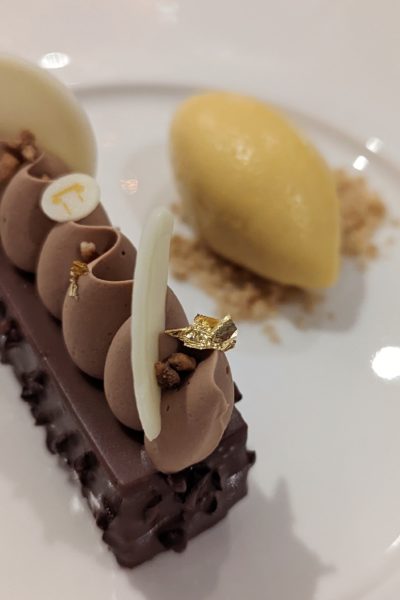 close up of a rectangular chocolate cake topped with chocolate moose and white chocolate circles next to a scoop of yellow ice cream on a white plate