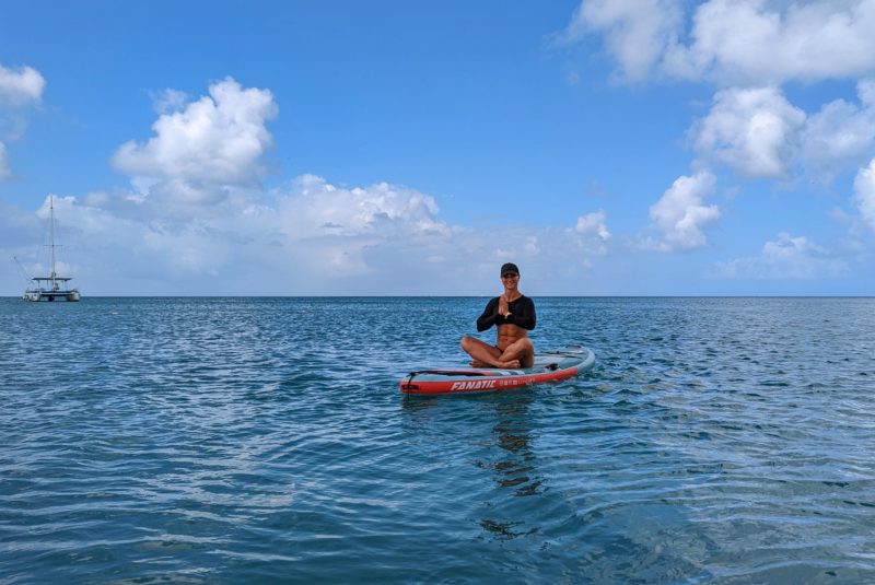 a blonde lady with very tanned white skin sitting crossed legged on a paddleboard on a calm blue sea with her palms pressed together in front of her chest. she is wearing a black long sleeved rash vest and black cap and smiling. there is a catamaran in the distance on the left. SUP yoga in aruba