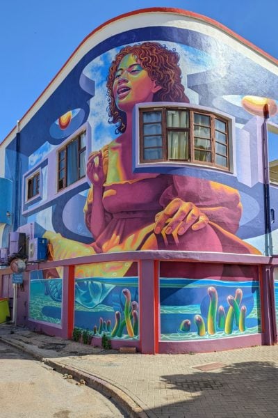large painted mural across two floors of a building in San Nicolas Aruba with a painting of a woman in a pink dress against a blue background