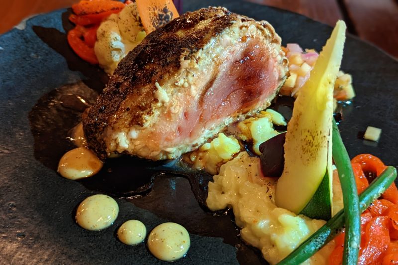 a black plate of food with a seared fillet of tuna still pink in the middle surrounded by several dots of yellow sauce and some vegetables