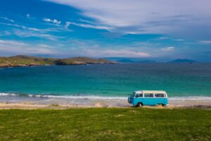 7 of the Best Places to go in a Campervan in the UK blue VW campervan driving on a road next to the sea in soctalnd with some low green cliffs in the distance behind on a very sunny day with blue sky above. best places to go in a campervan UK.