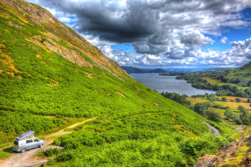 aerial shot of a large green hill with a dirt road in the bottom left corner where a grey campervan with a pop tent top is parked and in the distance on the right is a view of the blue lake at Ullswater in the Lake District in England. best places to go in a campervan UK.