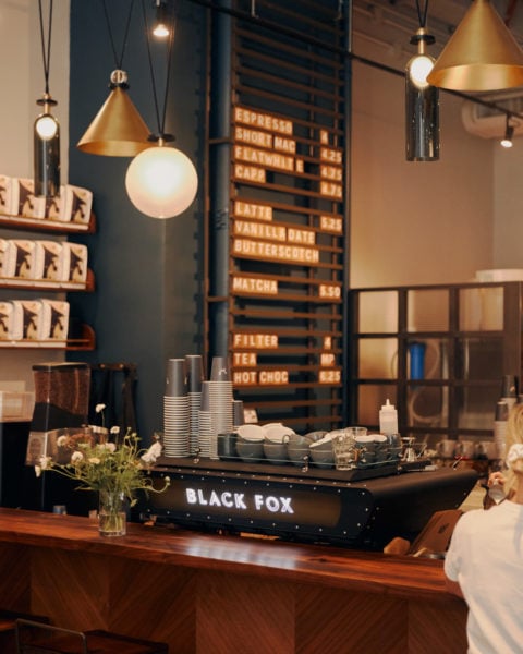 counter in a coffee shop made from dark reddish wood with a black sign on top reading Black Fox and the menu on the wall in white tiled letters slightly out of focus in the background