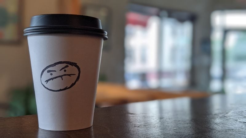 white paper coffee cup with a grumpy face logo on the side on top of a brown wooden counter top with the interior of an NYC coffee shop out of focus in the background