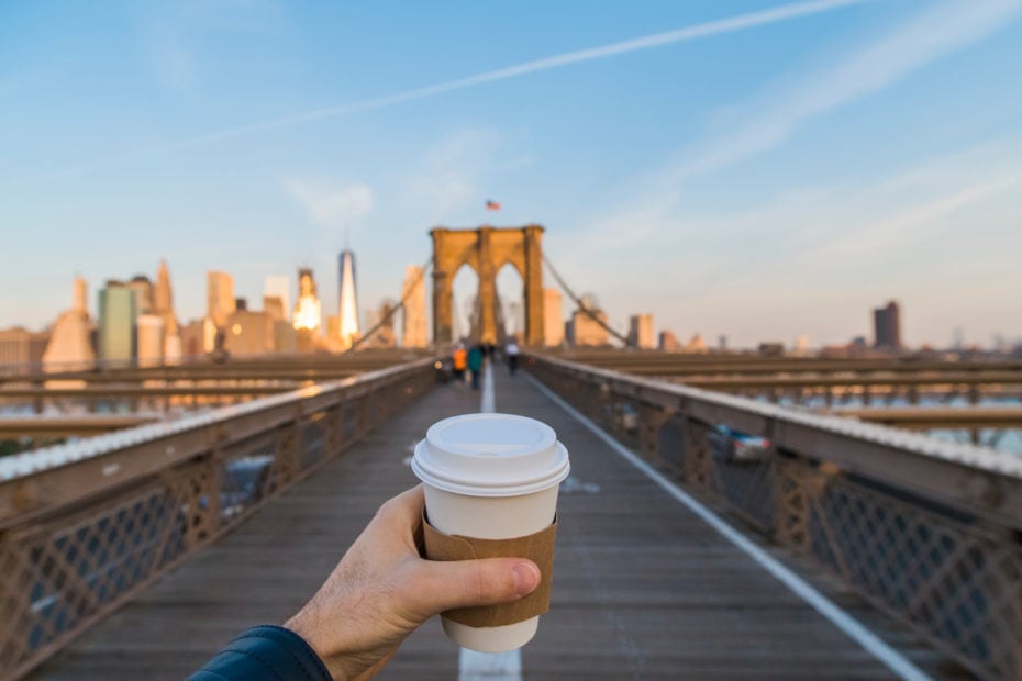 man's hand holding a white paper coffee cup with cardboard ring in front of the brooklyn bridge with the new york city skyline out of focus behind on a sunny day with a clear blue sky. Best coffee shops in NYC.