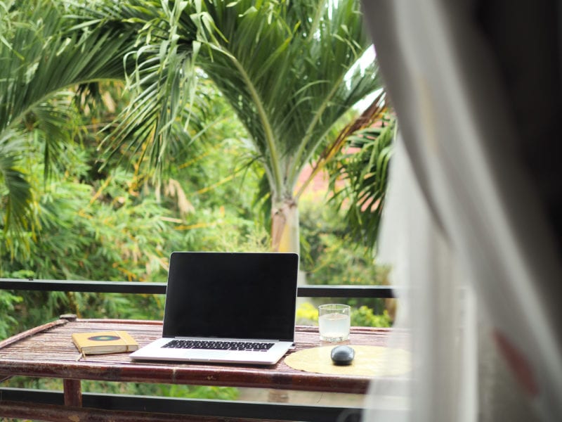 a wooden desk on a balcony with a laptop and notebook on it in front of a thick forest of palm trees and other exotic greenery with white curtains out of focus in the foreground on the right - how to make money from a travel blog