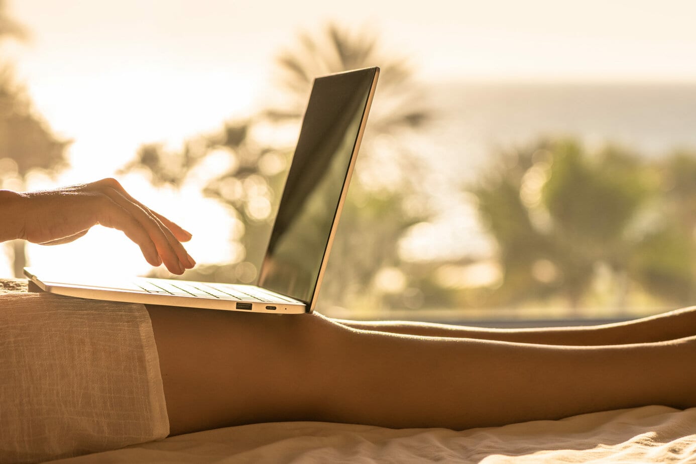 Close up of a woman's bare legs with a white skirt with laptop sitting on the bed in bedroom opposite panoramic window with view on palm tree sea beach at sunset - how to make money from a travel blog