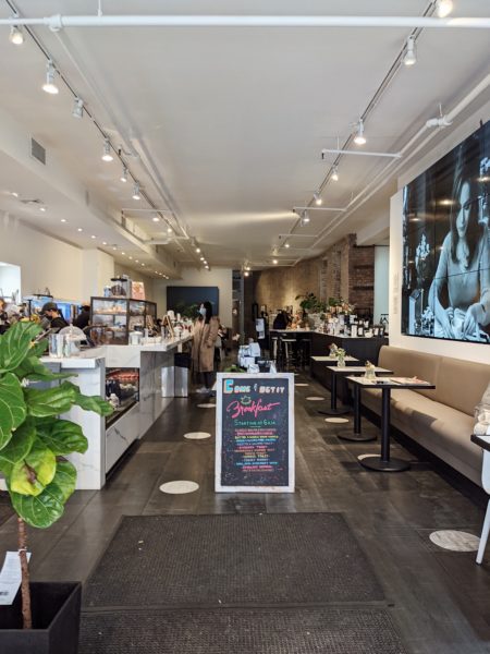 interior of a coffee shop in NYC with a grey floor and white ceiling and several wooden tables along a cream leather bench