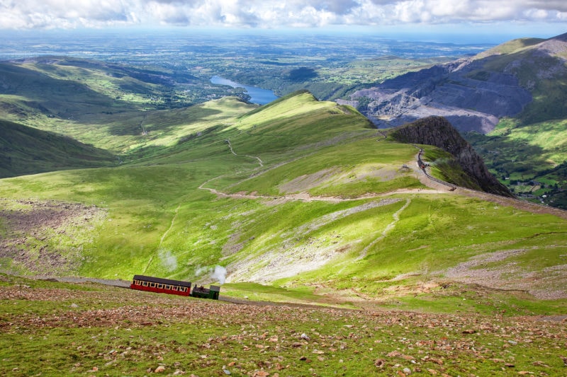 a small green steam train pulling a single red carriage seen very small against a landscape of green hills with a blue lake in the distance on a sunny day - things to do in llanberis