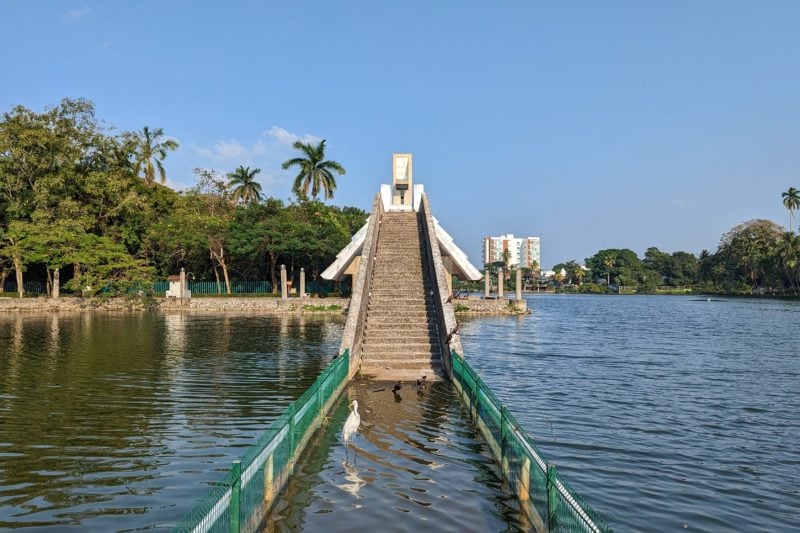 a blue lake on a very sunny day with blue sky above. there are stone steps leading towards a bridge over a section of the lake but the area in front of the steps is filled with water. a white Great Egret is standing in the water in front of the steps. 
