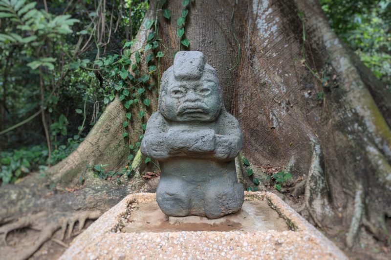 small stone sculpture  of a short animal like figure holding a rectangular object carved by a mesoamerican civilisation in front of a large tree at parque museo la venta