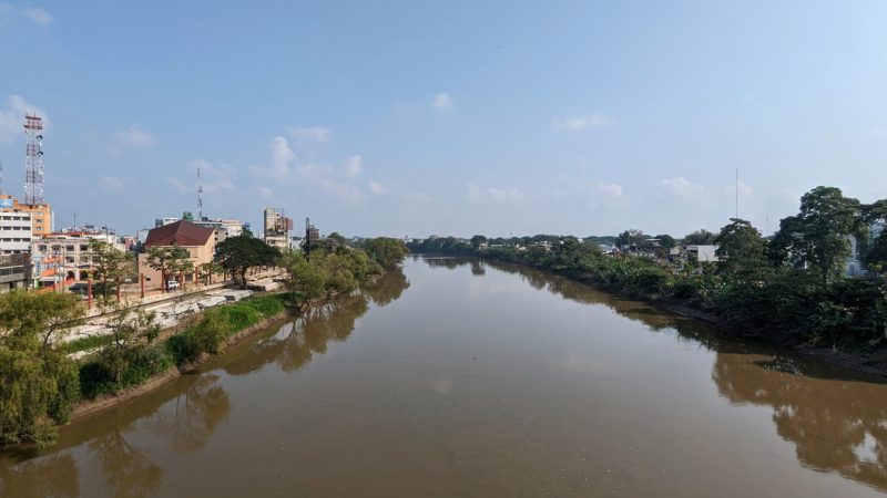 a brown coloured river viewed from a bridge on a sunny day with blue sk above. there are trees on the right bank of the river and the edge of villahermosa city visible on the left. 