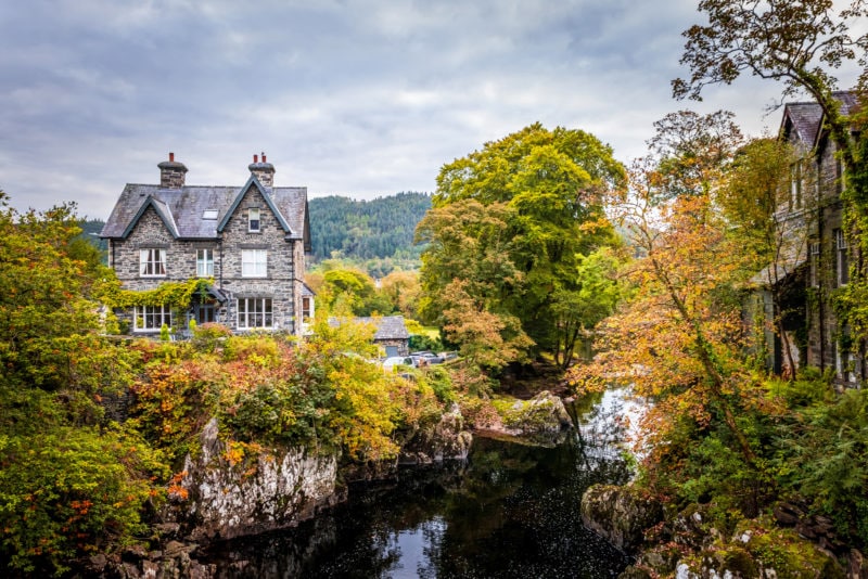 view of Betws-y-Coed in snowdonia wales on a grey day with overcast sky and lots of autumnal coloured trees. there is a grey stone house next to a river surrounded by trees. 