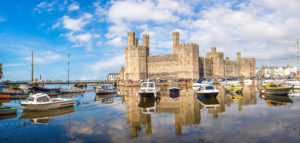 Caernarfon Castle in Wales, a large historic castle with multiple towers and battlements next to a tranquil body of water. Several boats are docked in the foreground, their reflections shimmering on the calm surface. The scene is picturesque with partly cloudy skies, patches of blue