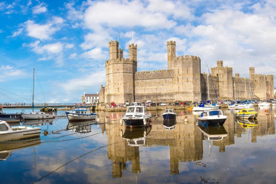 Caernarfon Castle in Wales, a large historic castle with multiple towers and battlements next to a tranquil body of water. Several boats are docked in the foreground, their reflections shimmering on the calm surface. The scene is picturesque with partly cloudy skies, patches of blue