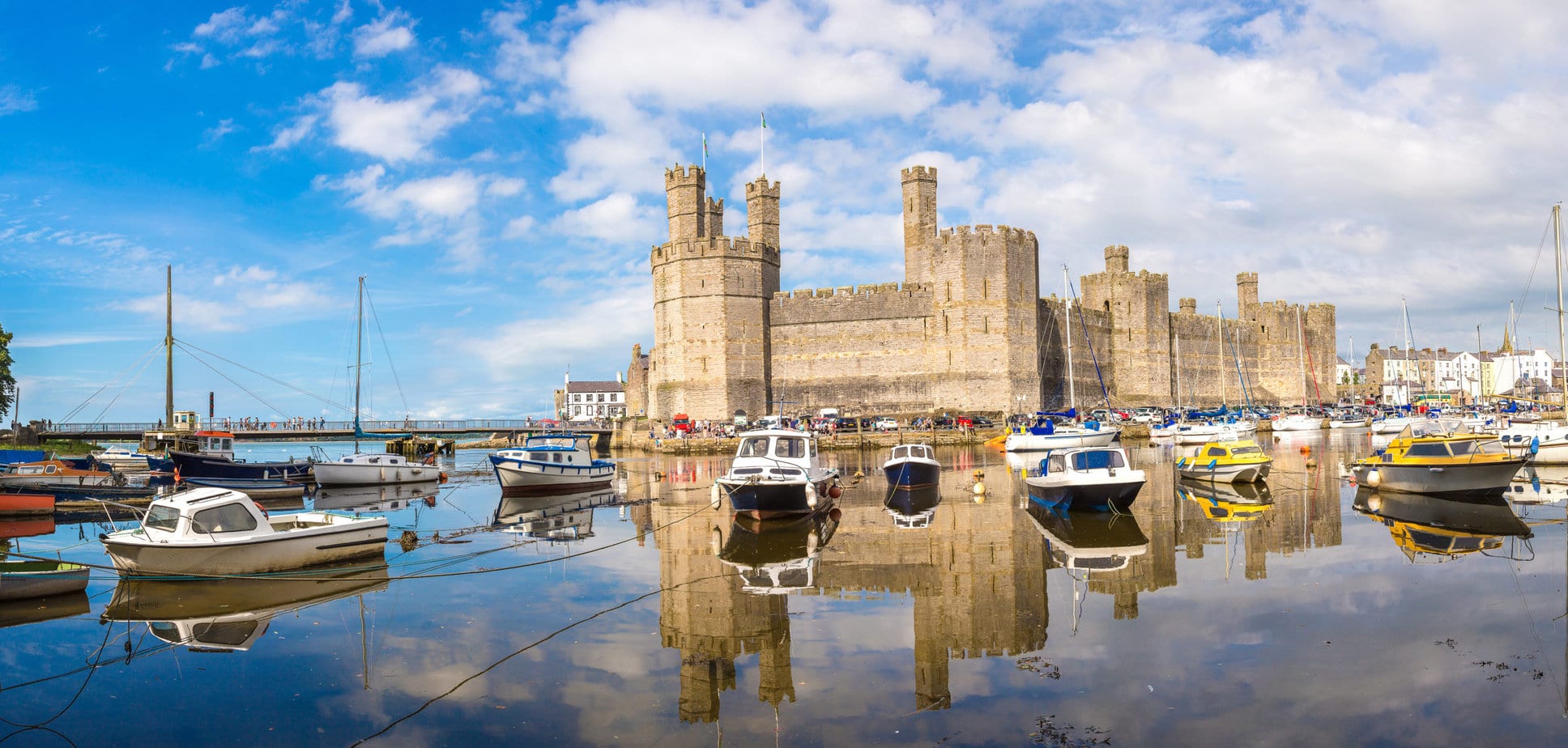 Caernarfon Castle in Wales, a large historic castle with multiple towers and battlements next to a tranquil body of water. Several boats are docked in the foreground, their reflections shimmering on the calm surface. The scene is picturesque with partly cloudy skies, patches of blue