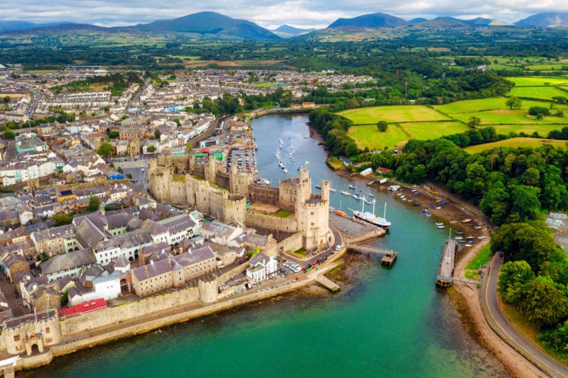 Aerial view of historical town Caernarfon in Wales, with a castle next to a very blue river. the town is on the left and on the right side of the riverbank are green fields and woodland. there are mountains in the background. 