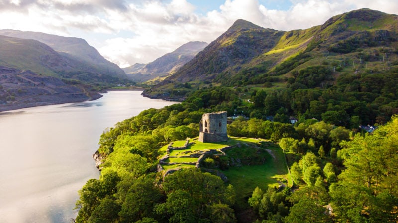 aerial shot of a small round stone castle turret on a green grassy hilltop surrounded by trees next to a very still blue grey lake with mountains in the background - Dolbadarn Castle in Llanberis Wales