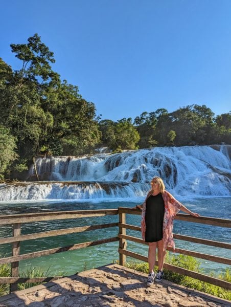 Emily wearing a black dress black converse and an orange shawl with gold sunglasses and her blonde hair down, standing in front of a pool of turquoise water with a small waterfall behind that on a very sunny day with blue sky overhead