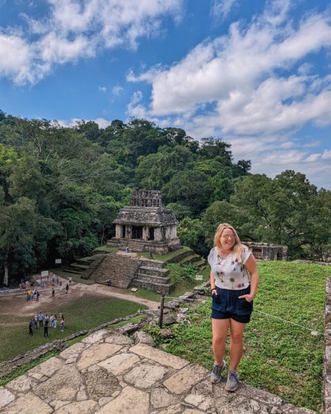 Emily wearing dark blue denim shorts and a white t shirt and gold sunglasses with her hands in her pockets bending forwards slightly laughing. she is standing on a stone step with a grassy ledge behind her and below her is a grey stone mayan pyramid at Palenque ruins in Mexico on a sunny day with blue sky overhead.