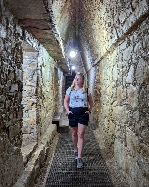 emily wearing dark denim shorts and a white t shirt with green palm trees on it walking towards the camera with her hands in her pockets with sunglasses on her head and her blonde hair loose. she is walking inside a narrow stone corridor with a few electric lights overhead inside a mayan pyramid at palenque