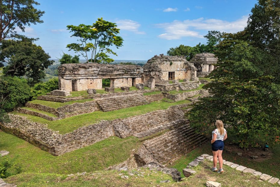 landscape at Palenque with ruins of a grey stone Mayan temple on a grassy hill surrounded by lush green trees. In the foreground is solo female travel blogger Emily Luxton wearing dark blue denim shorts and a white t-shirt standing on the edge of a hill looking out towards the ruins.