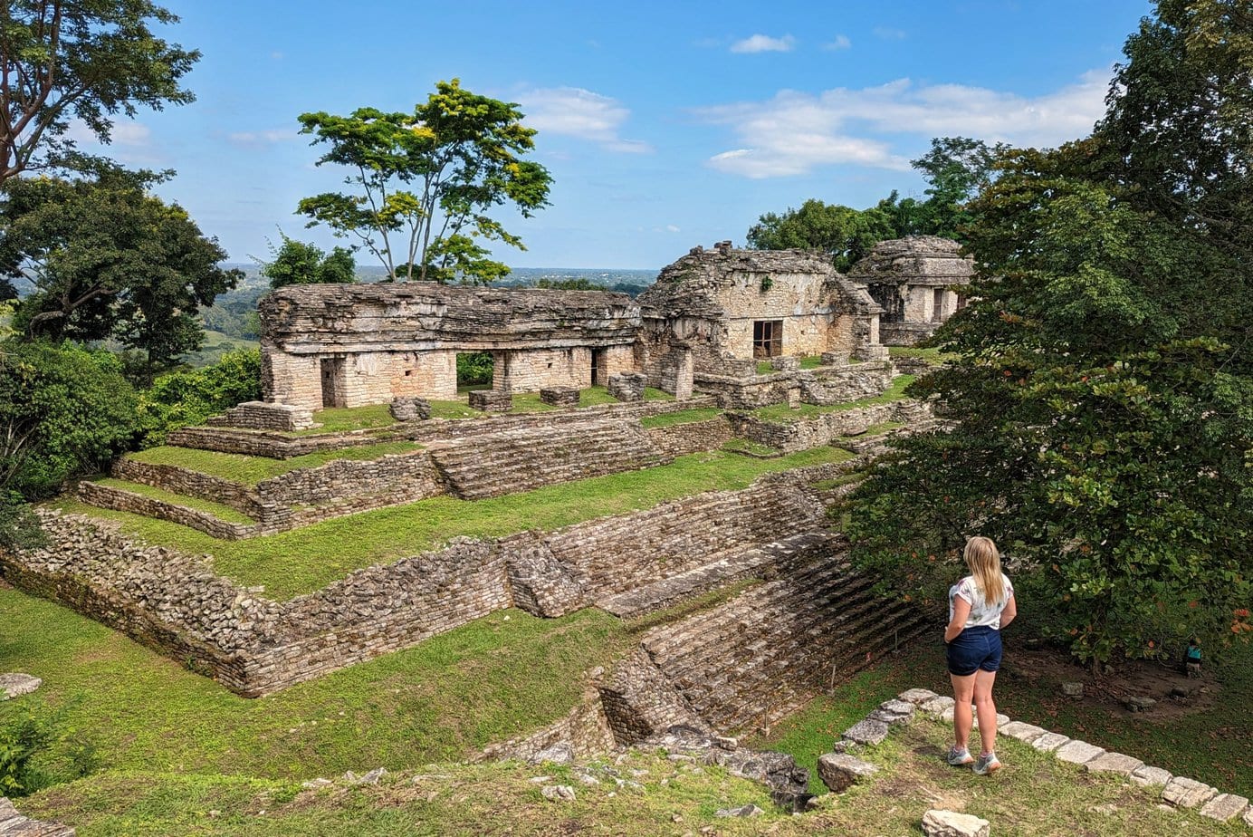 landscape at Palenque with ruins of a grey stone Mayan temple on a grassy hill surrounded by lush green trees. In the foreground is solo female travel blogger Emily Luxton wearing dark blue denim shorts and a white t-shirt standing on the edge of a hill looking out towards the ruins.