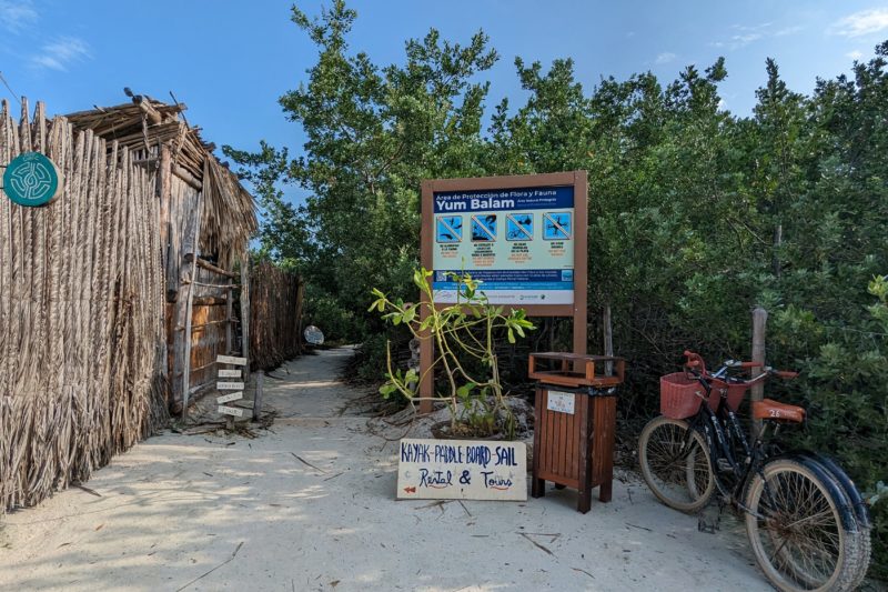 a sign for yum balam nature reserve in front of dense green bushes with a wooden fence on the left and a sandy path leading away into the reserve.