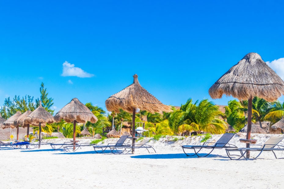 sun loungers and thatched-roof palapas style wooden umbrellas on a white sandy beach in holbox with palm trees and tropical foliage behind on a bright sunny day with vivid blue sky