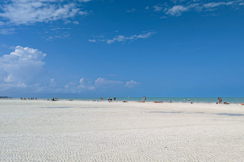 large empty expanse of white sand marked with ridges made by water with people in the distance along the waters edge on a very sunny day with clear blue sky overhead and a very thin strip of turquiose sea visible on the horizon