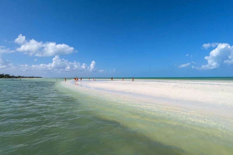 a sandbank made of bright white sand with water on both sides on a very sunny day with clear blue sky overhead. there are a few people walking on the sandbar in the distance. best beaches in holbox mexico. 