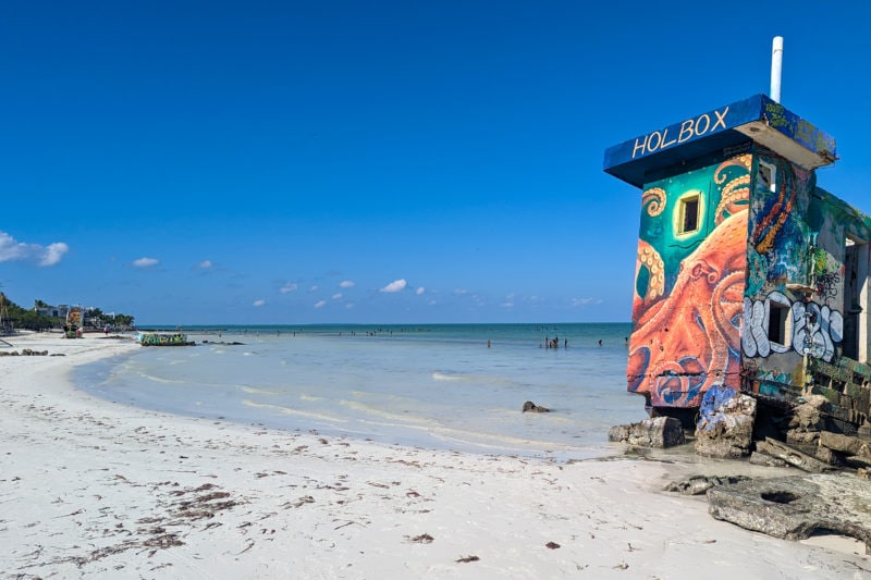 curved white sandy beach with shallow clear sea water on a very sunny day with blue sky and a ruined concrete building right on the waters edge painted with a brightly coloured mural of an orange octopus against a green background with the word Holbox printed at the top