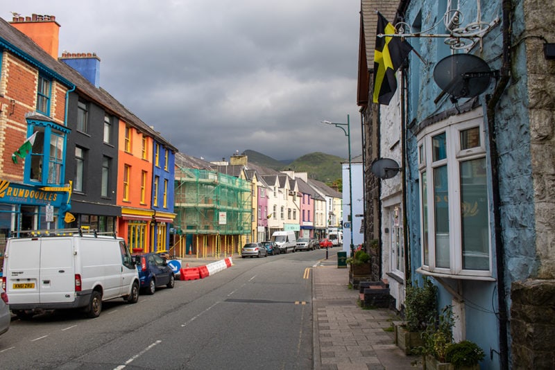 shot of Llanberis high street with colourful buildings and some scaffolding on either side of a road where a white van and black car is parked. there are mountains in the background and it is a very grey day with a cloudy sky. 