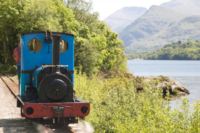 small blue steam train with a black face running along a track next to a lake on a sunny day in snowdonia wales