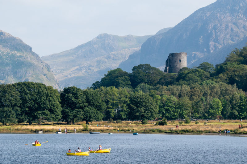 View of Dolbadarn Castle from across the water at Llyn Padarn with some small yellow rowboats on the blue lake and trees around the castle and mountains visible in the distance. Things to do in Llanberis Wales. 