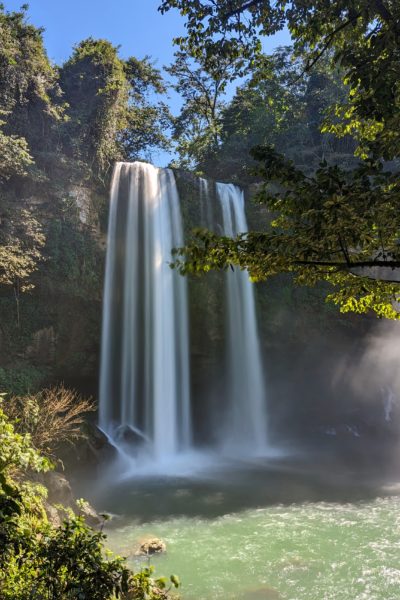 a tall waterfall taken with slow shutter so the water movement is blurred. the waterfall is emptying into a green pool surrounded by tocky cliffs and leafy trees.