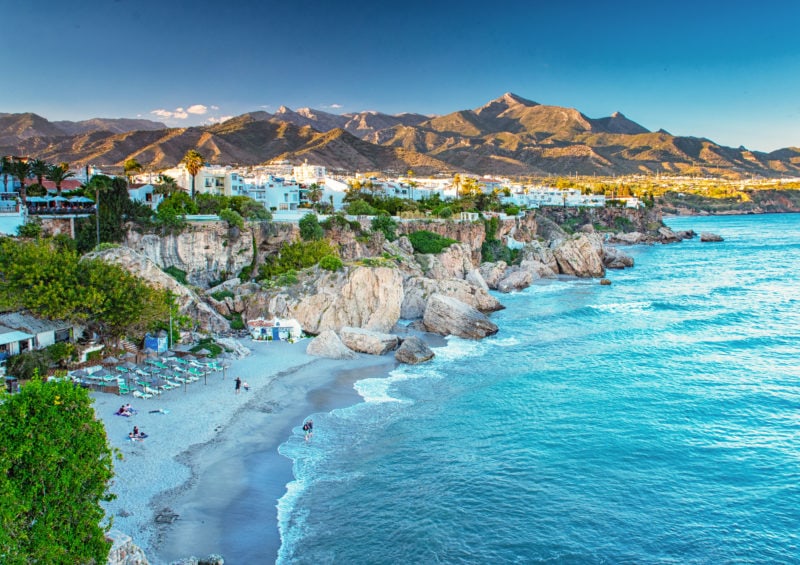 aerial shot of a white sandy beach and bright blue water with a white town behind and low sand coloured mountains behind that on a very sunny day in southern spain with blue sky overhead. Nerja is one of the best day trips from Malaga