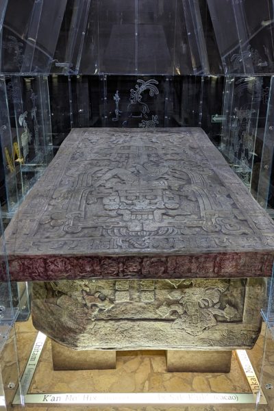 a large stone tomb covered with mayan carvings behind glass at a museum in palenque with black walls in the background