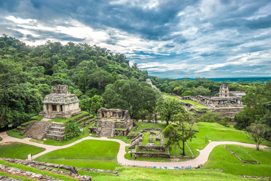 landscape with several grey stone pyramids and Mayan ruins dotted around a large green grassy area with dense forest to the left hand side of the image. How to get to Palenque in Chiapas Mexico.