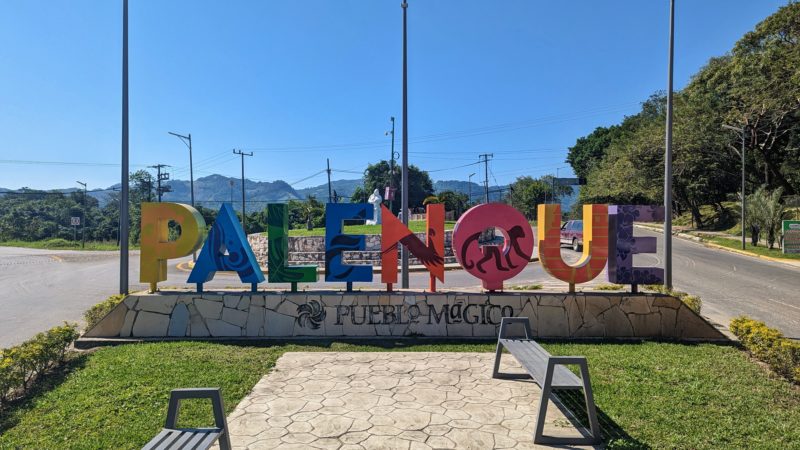 grassy area in between two roads with large sculpted letters in different colours of the rainbow spelling out the word Palenque with blue sky and mountains behind on a very sunny day