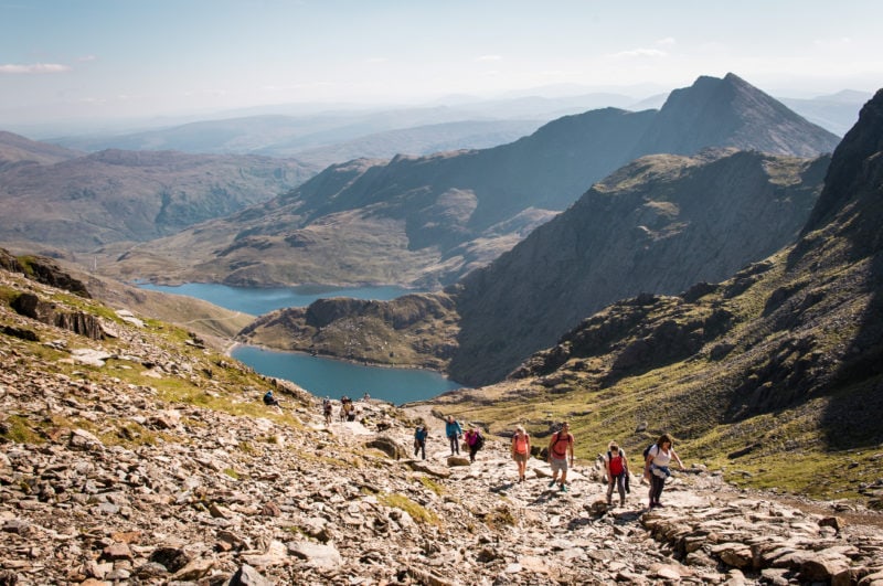 looking down a mountain with a line of people hiking up a stone path with two bright blue lakes in the background - climbing mount snowdon in wales on a clear day with a hazy blue sky overhead