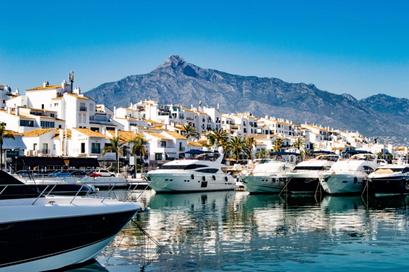 several yachts in a marina with bright blue water in front of a whitewashed spanish town with a mountain in the background and blue sky overhead. 