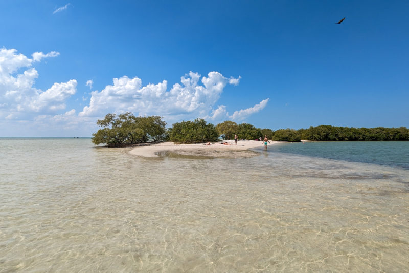 tiny beach with a few green trees on the end of a strip of land viewed from the water with the sea on one side and the blue water of a lagoon on the other and shallow clear sea in front on a very sunny day with blue sky overhead and a few fluffy clouds. 
