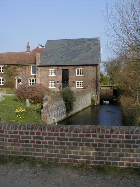 view of a beige stone cottage with grey roof next to a small river with a red brick wall in the foreground on a dull day in spring. there is a lawn in front of the cottage with daffodils on it. Redbournbury Mill in St Albans England