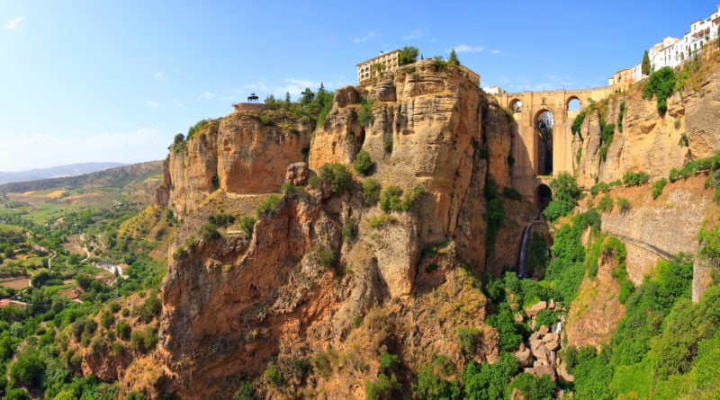 shot of a beige coloured building a tthe top of an orange cliff face with a large stone bridge above a waterfall connecting the cliff to another cliff. The Ronda is one of the best day trips from Malaga. 