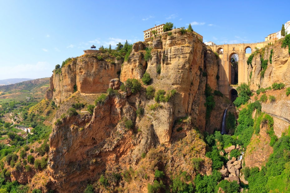shot of a beige coloured building a tthe top of an orange cliff face with a large stone bridge above a waterfall connecting the cliff to another cliff. The Ronda is one of the best day trips from Malaga.